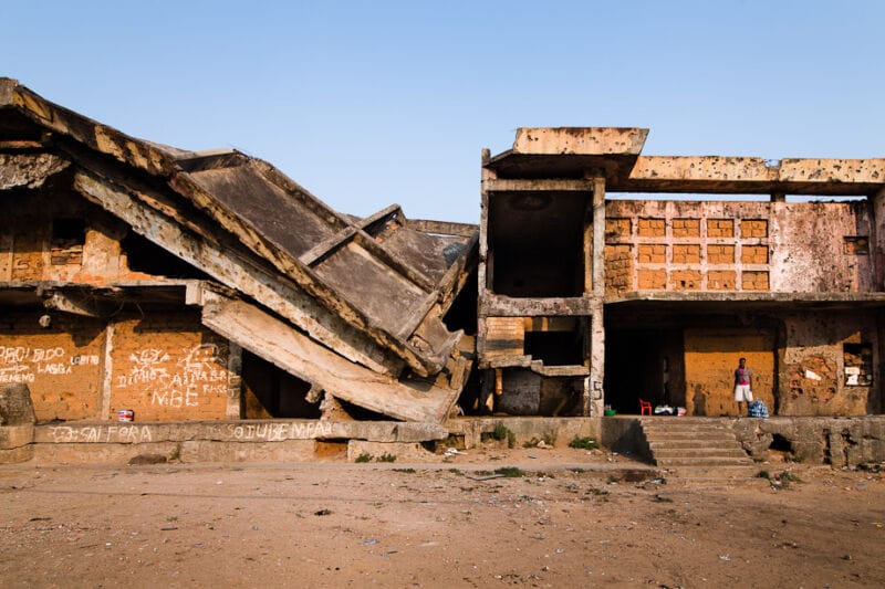 Edificio dañado por la guerra civil en Kuito, Angola, con los forjados de hormigón colapsados y la fachada marcada por impactos de metralla; una persona permanece de pie en una de las estancias abiertas